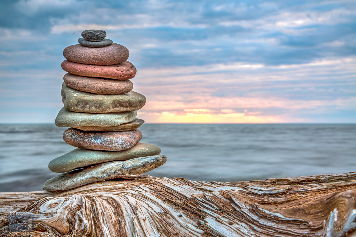 Photograph of a cairn (stack of rocks) on a driftwood log with Lake Superior and the sunset behind.